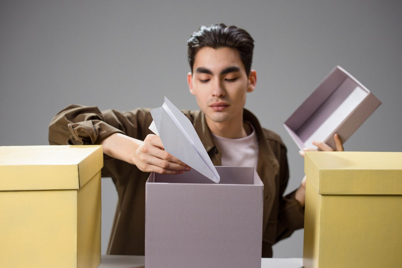 Asian man with dark hair holding a paper airplane above a large gift box indoors.